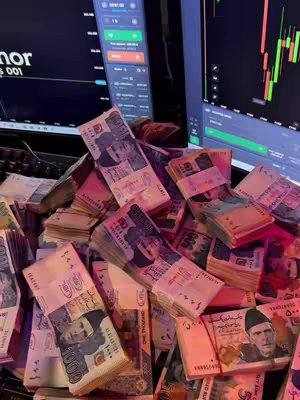 A close-up, top-down shot of a large pile of Pakistani rupee banknotes, with stacks and loose notes filling the frame. Two computer monitors displaying trading graphs are visible in the background, creating a contrast between the currency and financial analysis.
