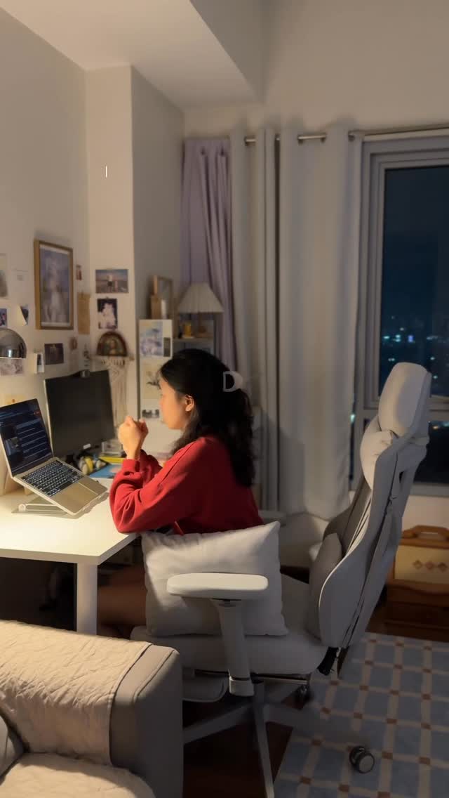 A cozy, dimly lit room with a woman seated at a white desk, working on a laptop. The lighting appears to be warm, casting shadows and highlighting the subject. The woman is wearing a red sweater, adding a strong visual contrast. In the background, a window offers a view of a cityscape at night. The composition includes a computer monitor, a comfortable chair, and personal items, creating a lived-in, relatable atmosphere. The overall mood is calm and focused.