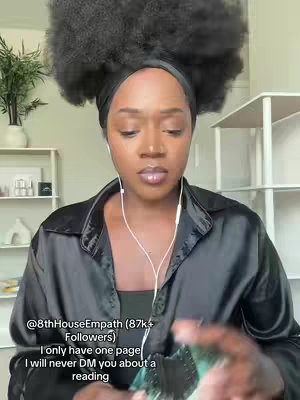 A woman with a large afro and a black headscarf is looking directly at the camera. She is wearing a black button-up shirt over a black top and has earbuds in. The background includes light-colored walls, shelves, and greenery.