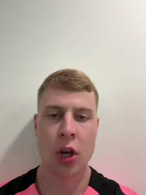 A medium shot features a young man's face centered against a plain white background. He has short, light brown hair. He is wearing a black shirt with a pink section around the shoulders and upper chest. The man's mouth is open as if he is speaking.