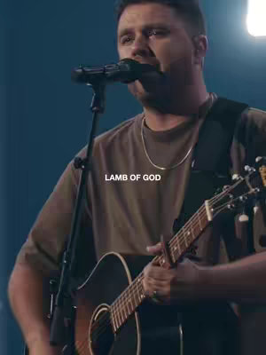 The image is a medium shot of a man playing an acoustic guitar and singing into a microphone. He is wearing a brown t-shirt and a gold chain. Below the man, overlaid in white text, is 'LAMB OF GOD'. The background is dark, suggesting a stage setting during a performance.