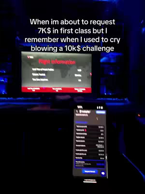 The image shows a dimly lit, dark interior. A TV screen, displaying flight information, is in the background. In the foreground, a smartphone is visible, showing a screen with detailed financial data. The text overlay provides context regarding first-class travel and financial challenges in the past.