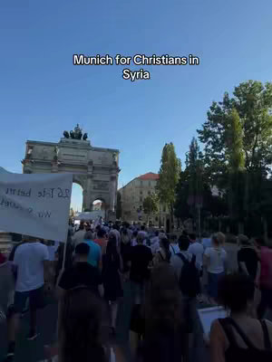 A crowd of people walking down a street with a large archway in the background. The sky is clear blue. Overlayed text states 'Munich for Christians in Syria'.