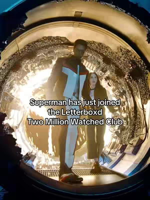 The image features a wide-angle shot of two individuals standing inside a circular enclosure. The background is filled with a complex, metallic structure illuminated by bright lights, creating a sense of depth and a futuristic or industrial setting. In the foreground are two figures: a tall, slender man in a suit of red, white, and blue colors, and a woman next to him wearing coat. The composition utilizes a low angle, making the figures appear prominent against the detailed background. The colors are vivid.