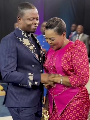 A man in a navy blue suit with a floral patterned tie stands with a woman in a pink patterned dress. They are looking at each other with a slightly downward gaze, holding hands.