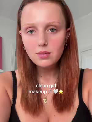 A close-up shot of a young woman with light skin and auburn hair that falls to her shoulders. She has fair eyebrows and is wearing a black top. The image has overlay text that says "clean girl makeup."