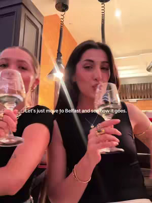 Two young women are holding glasses of wine in a restaurant setting. The women have their hands intertwined and are smiling. The backdrop features a warm-toned wall with overhead lighting.