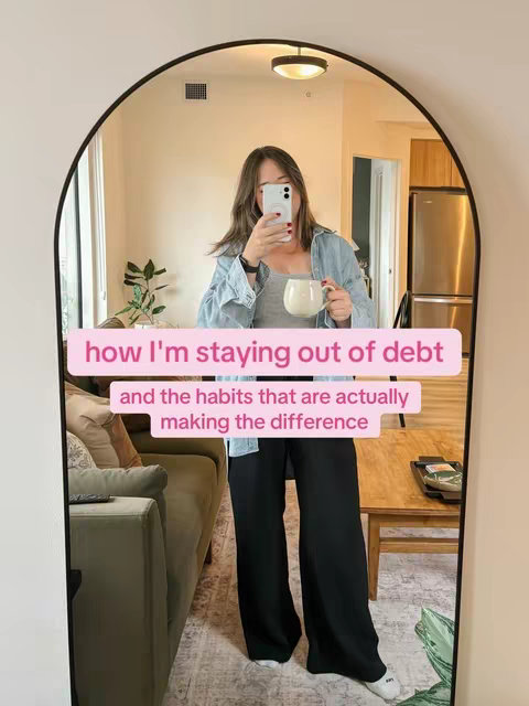 A woman is reflected in a large arched mirror. She is taking a selfie with her phone, holding a mug. She's wearing a light blue denim shirt over a gray top and black pants. The background shows a living room with a sofa, wooden coffee table, and a glimpse of a kitchen. Text overlays give context to the video's theme.
