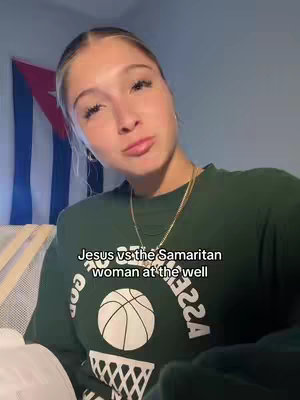 A young woman with blonde hair is filming herself from the chest up. She wears a dark green sweatshirt with a basketball graphic that says "ASS
22A" around the bottom of the circle. Her expression is neutral, looking slightly up toward the top left corner. The background includes a flag and a wall.
