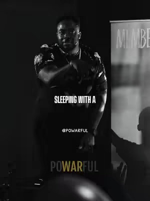 Black and white image with a man in a floral shirt standing in front of a dark background. White text overlays the image with the words 'SLEEPING WITH A'. There's also text that reads '@POWARFUL' and 'POWARFUL' below. The composition is shadowed, with the man's pose and expression conveying intensity.