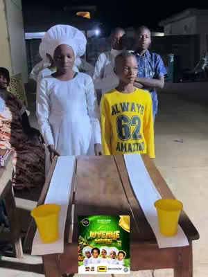 A group of children in white and yellow attire stand behind a table with two yellow cups and a poster that says 'JUVENILE HARVEST'.