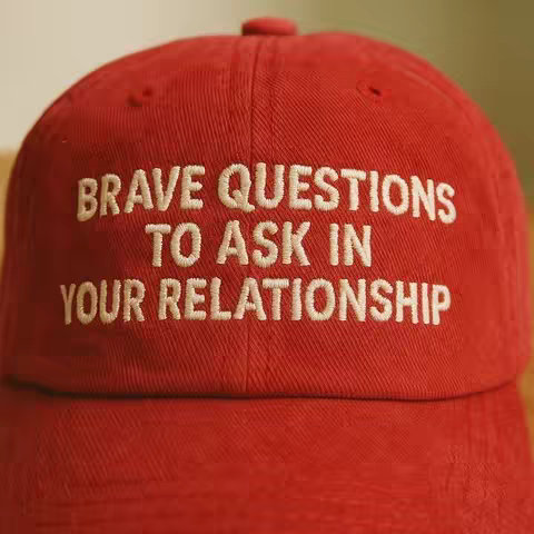 The image is a close-up of a red baseball cap. The cap has white, embroidered text. The text reads 'BRAVE QUESTIONS TO ASK IN YOUR RELATIONSHIP.' The background is blurred, suggesting a shallow depth of field.