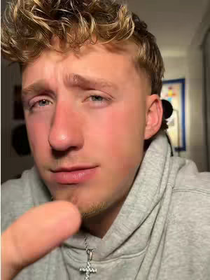A close-up shot of a young man wearing a gray hoodie. He has short, styled blonde hair and is looking towards the camera with a slight expression. Part of his finger is in the foreground pointing towards the camera. A silver cross necklace hangs around his neck. The lighting suggests an indoor setting, and a painting is visible in the background.