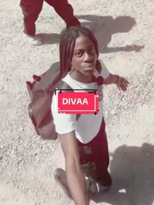 A person kneels on a sandy surface, looking towards the camera. They wear a white shirt, dark pants, and a backpack. A red and white graphic, containing the word 'DIVAA,' is superimposed near their chest. Another person’s legs and feet are visible in the background.