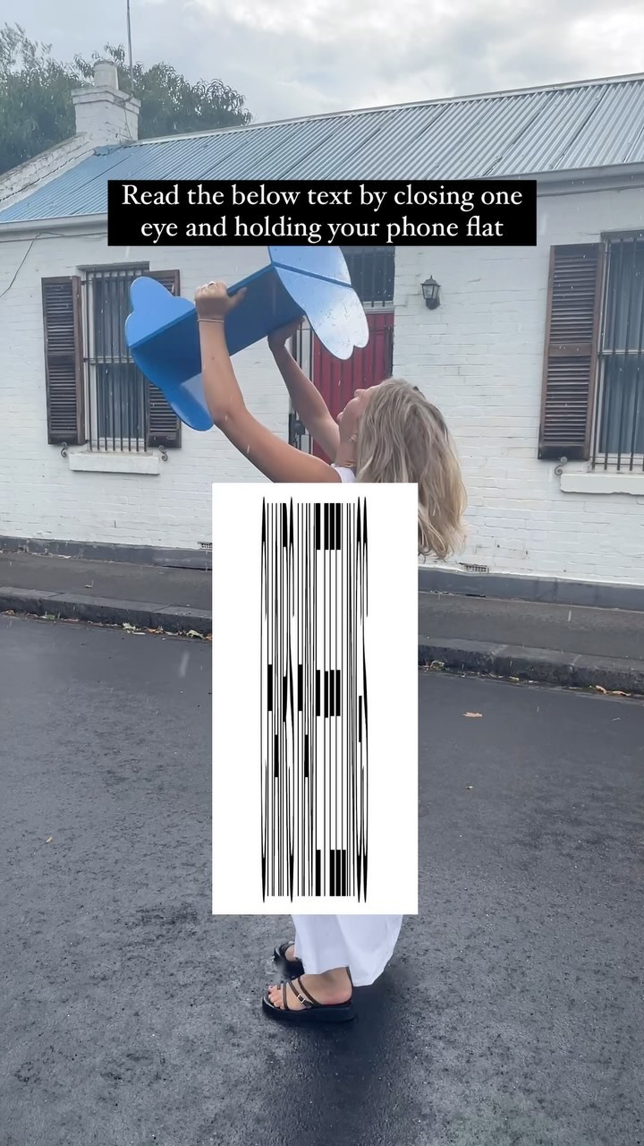 A woman is holding a blue object over her head on a rainy street. Beneath her is a white rectangle with a black and white barcode-like image. The background shows a building and a wet street.