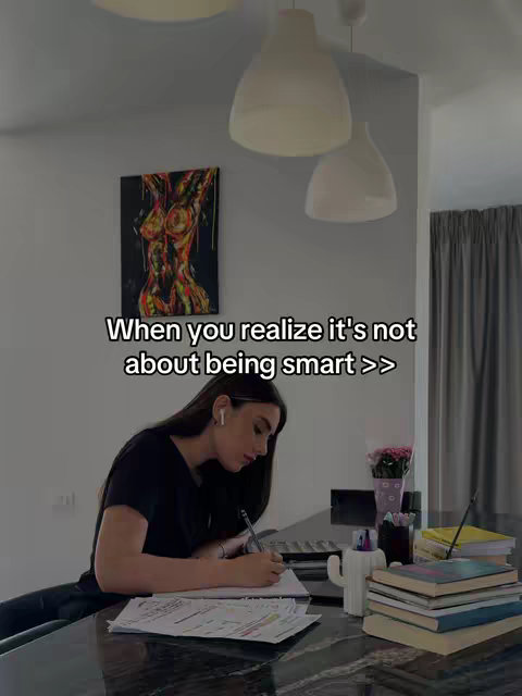 A woman sits at a desk, writing on paper. In the background is an abstract painting of a torso, a wall, and a window. The image is softly lit, with a muted color palette.
