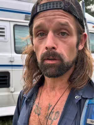 A man with a beard and a backwards cap stands in front of a van, looking at the camera. He has long, wavy hair and is wearing a blue collared work shirt. Tattoos are visible on his chest.