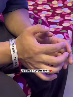 The image shows two hands clasped together, with the focus on the hands. The background is blurred. A silver bracelet is visible on one wrist, with the name "RAHUL" printed on it. Text overlayed at the bottom of the image reads, "You are an overthinker ❤️".