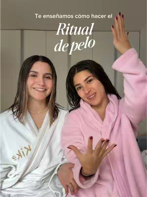 Two women stand side-by-side in a well-lit room, both smiling at the camera. The woman on the left is wearing a white robe with a gold embroidered logo. The woman on the right has a pink robe, holding her hand up and exposing her nails. Text overlays at the top read, in Spanish, "Te enseñamos cómo hacer el Ritual de pelo", which translates to "We'll show you how to do the Hair Ritual."