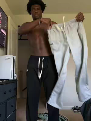 A shirtless Black man holding up two pairs of sweatpants in front of a neutral background. He's holding a gray pair with the right hand and a black pair with the left hand. The image is well-lit, showing details of both the man's physique and the fabric of the sweatpants. The overall composition suggests a product showcase or fashion-related content.