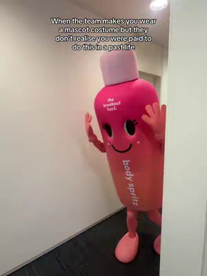 The cover image features a person in a pink and red mascot costume of a body spray bottle, standing in a hallway. The costume has a smiling face and the words "body spritz" printed on it. The background is a plain white wall and a dark floor.