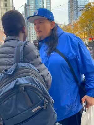 A man wearing a blue baseball cap and jacket is talking to someone off-camera. The background shows a cityscape and a second person with their back to the camera.