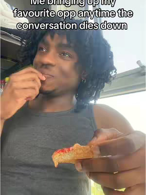 A selfie-style shot featuring a young Black man in a gray tank top, holding a piece of food, likely bread with red spread. He looks up slightly with a subtle smile, suggesting amusement or a playful expression.