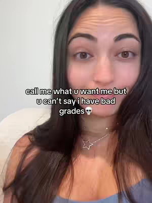 A selfie-style video cover image features a young woman with brown hair and dark eyes looking directly at the camera. The top part of the image is filled with her face, while the lower part shows her chest and a small star-shaped necklace. Overlaid in a sans-serif white font are the words, "call me what u want me but U can't say i have bad grades ", set against a blurred white background. A cartoon skull sits at the end of the word "grades". The image uses a soft, diffused lighting, giving the image a natural and intimate feel.