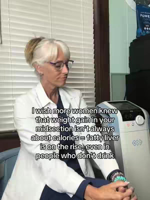 A woman with stylishly coiffed gray hair and glasses is centered in the image. She is wearing a white coat over a dark shirt and has a look of thoughtful concern. The background includes white blinds and a medical-looking appliance.