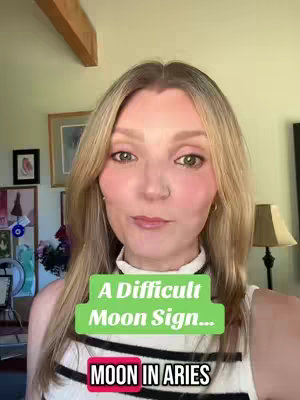 A woman with long blonde hair looks directly at the camera. She is wearing a white top. Overlayed are two text boxes, one green with the words 'A Difficult Moon Sign...' and another with a pink background labeled 'MOON IN ARIES'. The background setting includes indoor features like artwork on the wall and a lamp. The overall composition is a selfie shot.