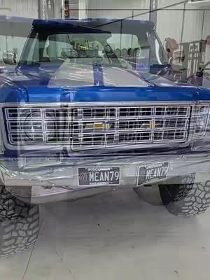 A front view of a blue Chevy truck is shown, angled slightly upwards. The focus is on the front, with a chrome grill and two license plates visible. Oversized tires are in view at the bottom. The truck is in a bright location, and the front of the vehicle is the main subject.