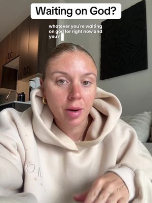 A woman in a beige hoodie is centered in a vertical frame. Above her, a white banner reads "Waiting on God?" Text below asks "whatever you're waiting on god for right now and you'r." Her blonde hair is pulled back, and she wears gold hoop earrings and a simple necklace. The background includes light-colored cabinets, a modern black abstract art piece, and a glimpse of a kitchen.