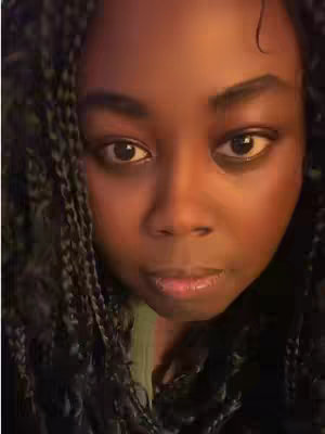 A close-up shot of a person's face, with the top of their head and shoulders visible. The person has braided hair and is looking directly at the camera. Their eyes are open and appear softly lit. The lighting on their face is warm, with both highlights and shadows.