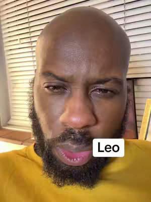 A close-up shot of a Black man with a shaved head and a dark beard. He appears to be looking slightly to the side with a somewhat quizzical or serious expression. His skin tone is dark, and he is wearing a yellow shirt. The background shows window blinds.