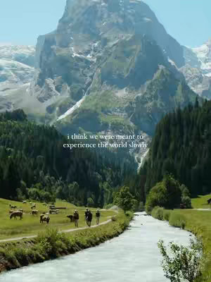 A scenic landscape featuring a mountain in the background. In the foreground, a river flows through a green valley where a few people are walking on the pathway. There are sheep grazing on the left.