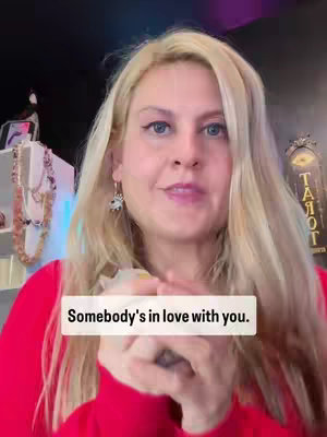 A woman with long blonde hair and pinkish-red shirt looks directly at the camera with a thoughtful expression, holding what appears to be a deck of cards in her hands. A white rectangular text box overlaid displays the text 'Somebody's in love with you'. The background is partially obscured by darkness lit with pink light with some decor visible to the left and a sign to the right.