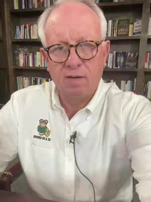 A man with gray hair and glasses is centered in the shot, wearing a white button-down shirt. Behind him is a bookshelf filled with books. The man is looking at the camera and appears to be speaking. A small logo is visible on his shirt. The lighting is natural and bright.