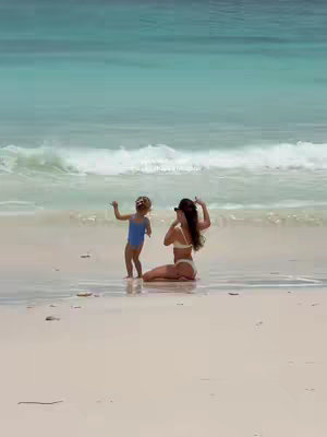 A sunny beach scene with a woman and a young child. The woman is kneeling in the shallow water, wearing a white bikini, and has her arms raised in the air. A young child, wearing a blue swimsuit, stands nearby with arms raised. The water is a vibrant turquoise, and the sand is light-colored. The sky is blue, with white waves in the background.