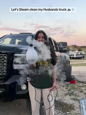 A woman stands in front of a large black truck. She's holding a white steam cleaner, with steam billowing out towards the camera. The background shows a rural setting at dusk.