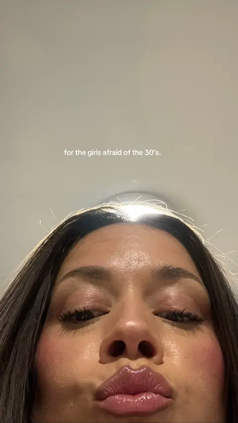 A close-up shot is focused on the lower part of a woman's face. Her lips, glossy and full, are puckered as if in a kiss. Her cheeks are flushed with a pink blush. Her eyes are partially visible with dark, long eyelashes. The camera angle is low, and the bright, plain background is a light color.