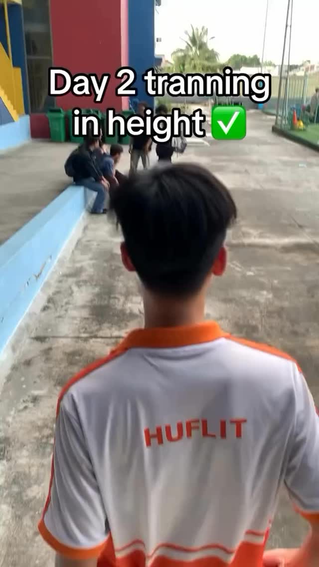 The image shows a young man from the back, facing away from the camera, walking down a path. He wears a white and orange shirt with the word 'HUFLIT' printed on the back. The foreground shows a pathway with a person walking towards the right. The background shows several people sitting and standing. Text overlays reads 'Day 2 tranning in height' with a green check mark.