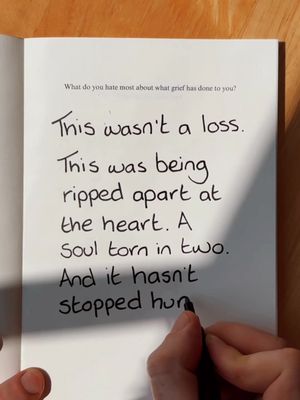 An open book on a wooden surface, showing a page filled with handwritten text. The text is in black ink and is the primary focus, with a hand holding a pen towards the bottom right of the frame as though writing. The top of the page includes the question "What do you hate most about what grief has done to you?" The lighting appears natural, creating shadows and highlighting the texture of the paper. The overall composition is simple and focused on the text.