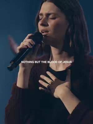 A woman is singing into a microphone while holding her hand over her heart. The image has a dark background, and the woman is wearing a dark top and maroon sweater.
