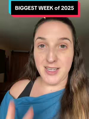 A woman with long brown hair is centered in the frame, looking directly at the viewer and speaking. Above her head is a rectangular text overlay with the words 'BIGGEST WEEK of 2025'. She’s wearing a sleeveless blue top over a black top. The background is a neutral-toned room with blurred details and a dark-brown wooden cabinet. The lighting appears to be natural, with the focus on the woman's face, which is framed by long, wavy hair.