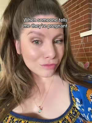 A selfie-style shot featuring a woman. She is looking towards the lower left, with a slightly uncertain or amused expression. She is wearing a blue patterned top and a gold necklace. A text overlay at the top says, 'When someone tells me they're pregnant.' The background includes a partially visible brick feature.