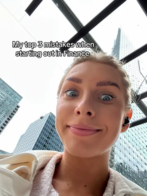 A selfie-style shot of a young woman against a background of modern architecture. The woman is positioned in the foreground, with her face prominently displayed, looking directly at the camera with a surprised expression. The background features tall buildings and a partially visible sky. The composition employs a low-angle perspective.