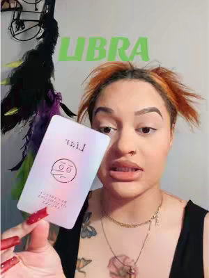 A woman with orange hair and light makeup holds a card, with the word "Libra" in the background. Also, there is a dark decoration on the top left and a blurred background.