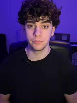 A young man with curly dark hair is centered in the frame, looking directly at the camera with a neutral expression. He wears a black t-shirt, and a small microphone is clipped to it. The background is blurred, but hints at an indoor setting with cool blue lighting and a musical instrument.