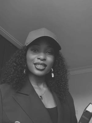 A grayscale photo with a woman as its subject. The woman has curly hair, earrings, and a necklace. She wears a baseball cap and a dark blazer over a black top. She has braces and appears to be smiling. The background is a simple, undefined setting.