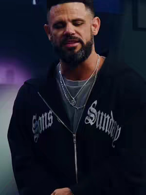 A medium shot features a man in a black hoodie, looking slightly downwards. The hoodie has a zipper and the text "Sons Sunday" in a stylized, white font across the chest. He wears a gray t-shirt underneath, and multiple silver necklaces are visible around his neck, one with a pendant. His hair is short and styled, and he has a trimmed beard. The background is a blurred mix of dark and purple hues.